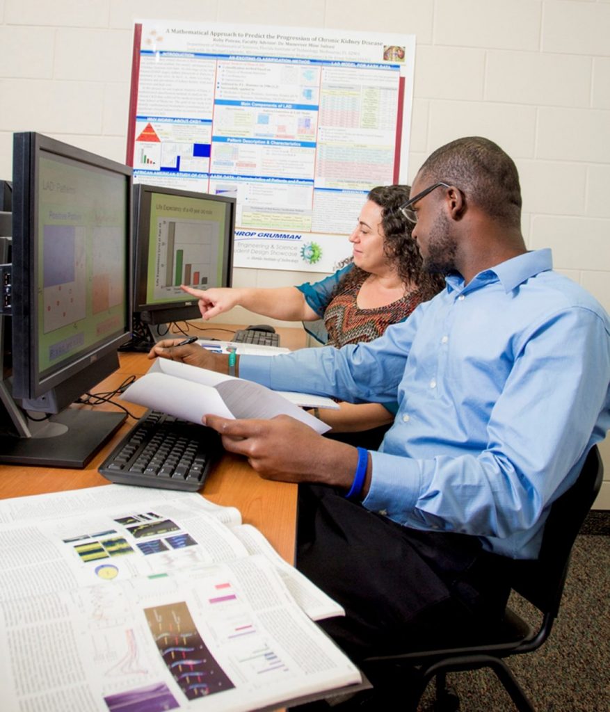 Teacher and student sit at computer with papers sprawled on the desk