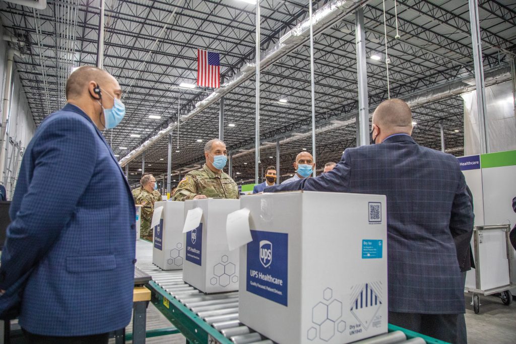 Two men in suits and some in military uniforms in a large warehouse with boxes of vaccines