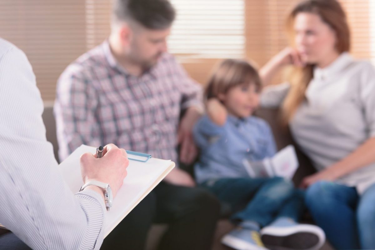 A family, blurred in the picture, sits on a coach as a person with a clipboard, likely a therapist, sits nearby.