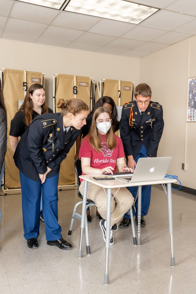 Florida Tech student sits at desk surrounded by high school students on a computer.