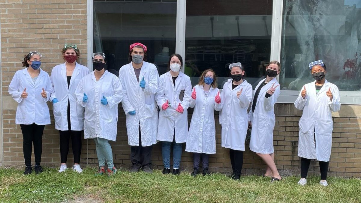 A row of nine students wearing lab coats poses for a picture outside