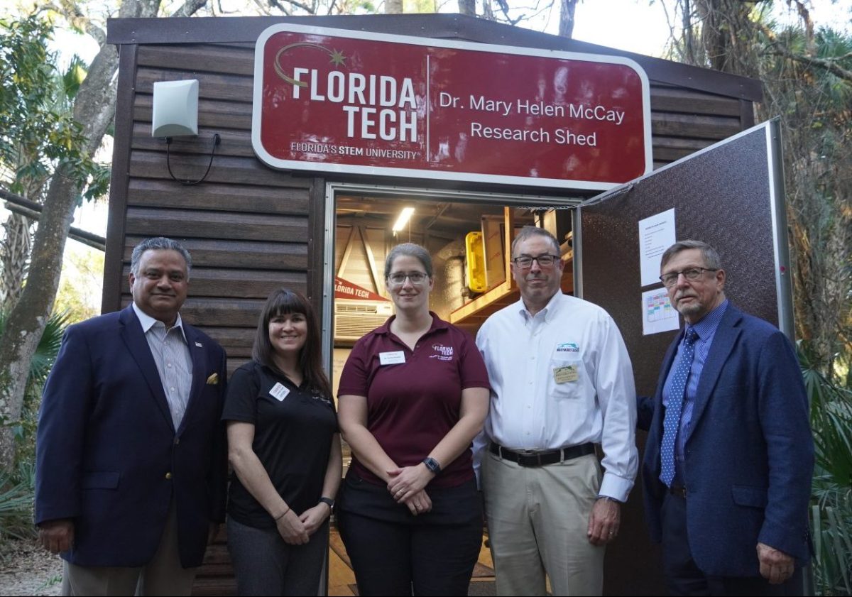 Five people stand oin front of a small, faux-wood sided shed.